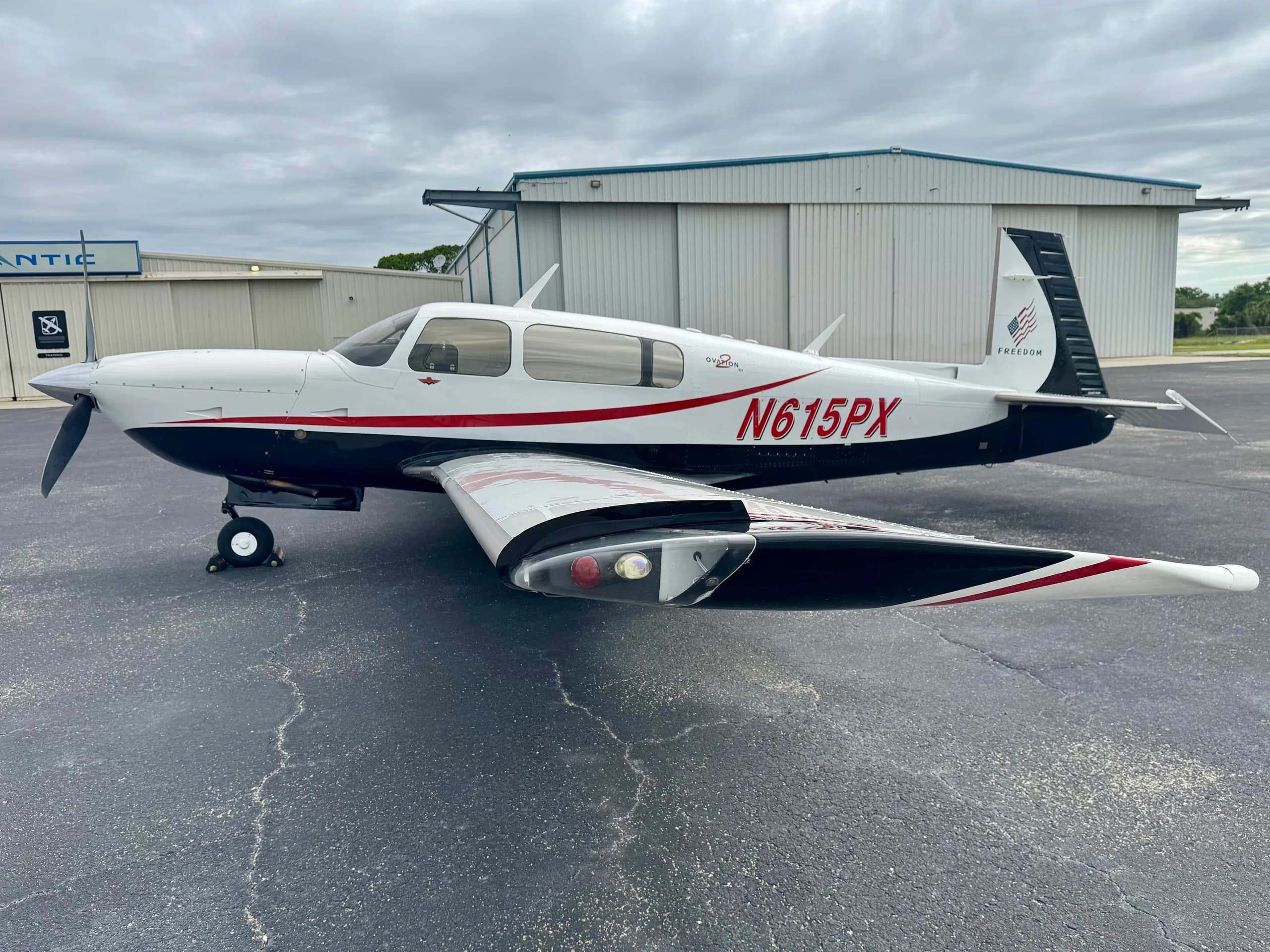 A white and black Mooney Ovation GX aircraft with red N-number N615PX parked on an asphalt ramp in front of a hangar.