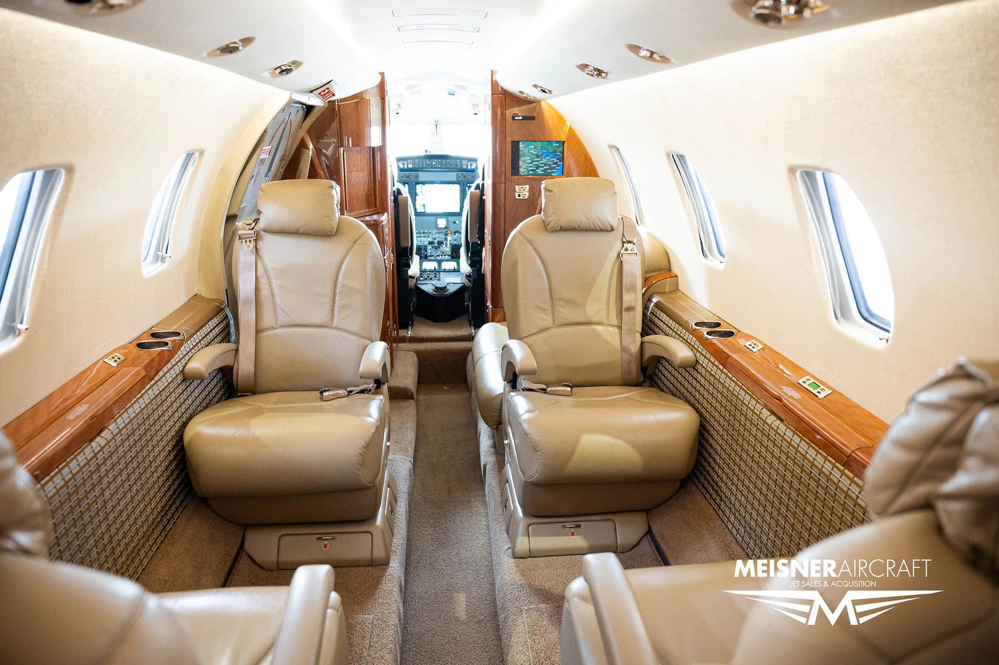 The luxurious interior cabin of a business jet with tan leather seats and wood accents, looking towards the cockpit, with a 'Meisner Aircraft' logo.