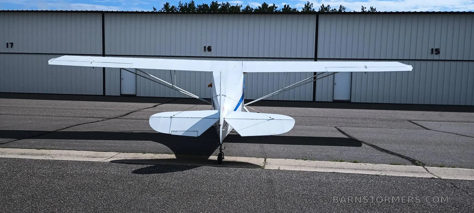 The rear view of a white single-engine taildragger aircraft parked on an asphalt ramp in front of hangars.