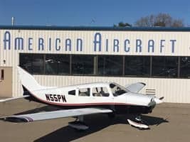 A white single-engine propeller aircraft with black and red trim is parked in front of a building labeled "AMERICAN AIRCRAFT".