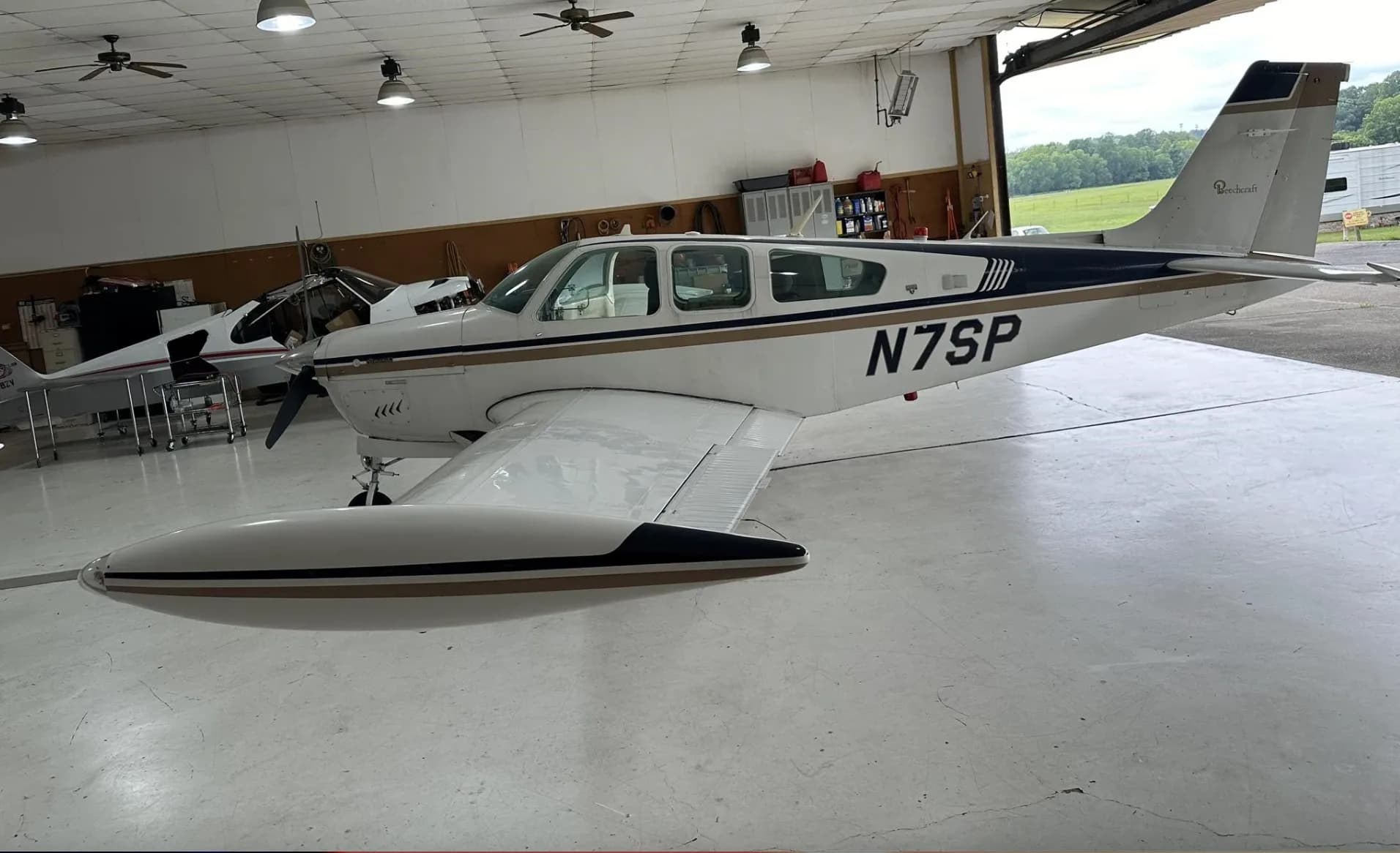 A white and gold Beechcraft Bonanza N7SP with tip tanks, parked in a hangar, viewed from the side.