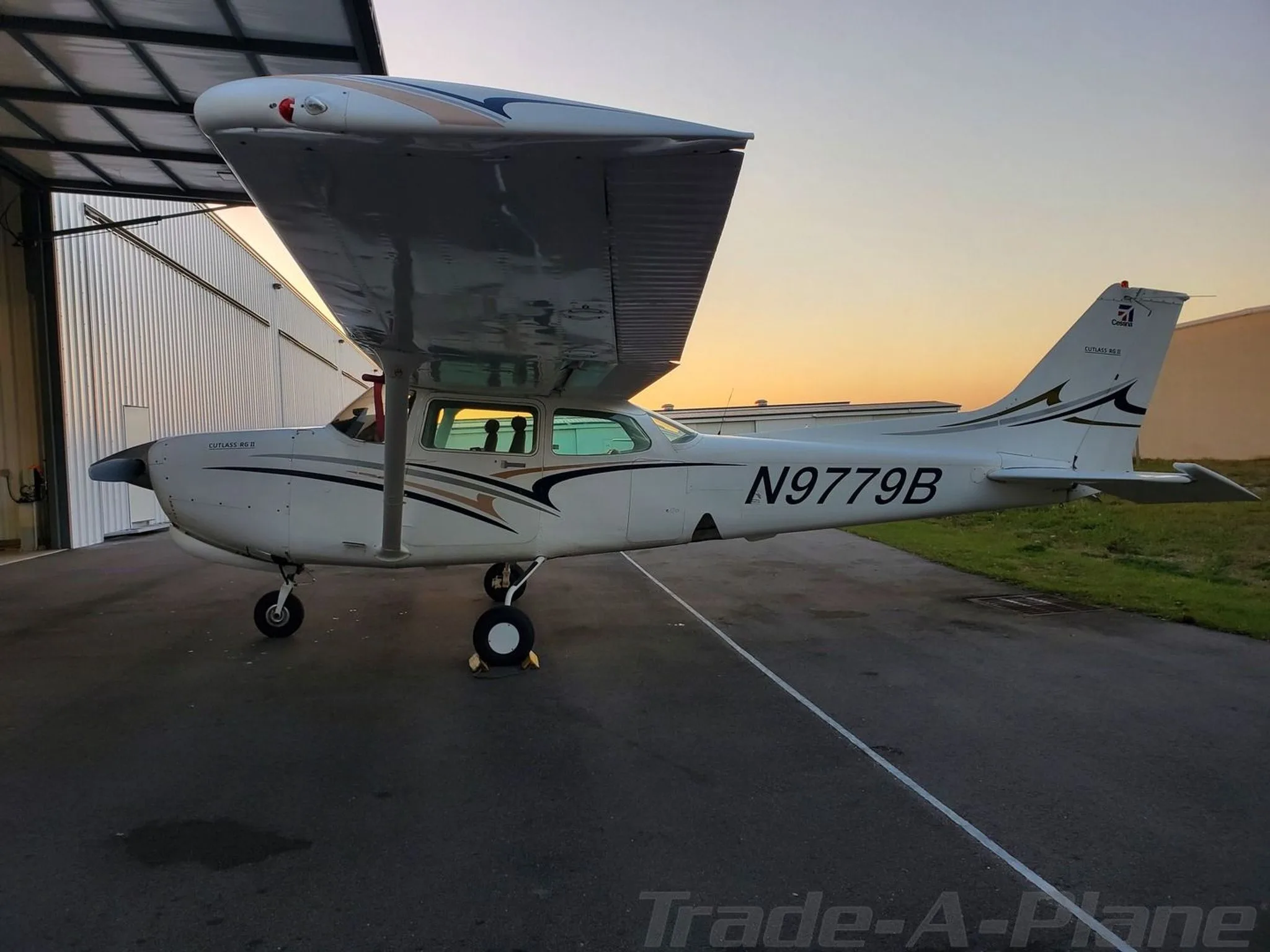 A white Cessna Cutlass RG II (N9779B) with black and gold stripes parked outside a hangar at sunset.
