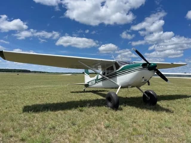 A white and green single-engine propeller aircraft with large tires, registration N82266, parked on a grassy field under a blue sky with clouds.