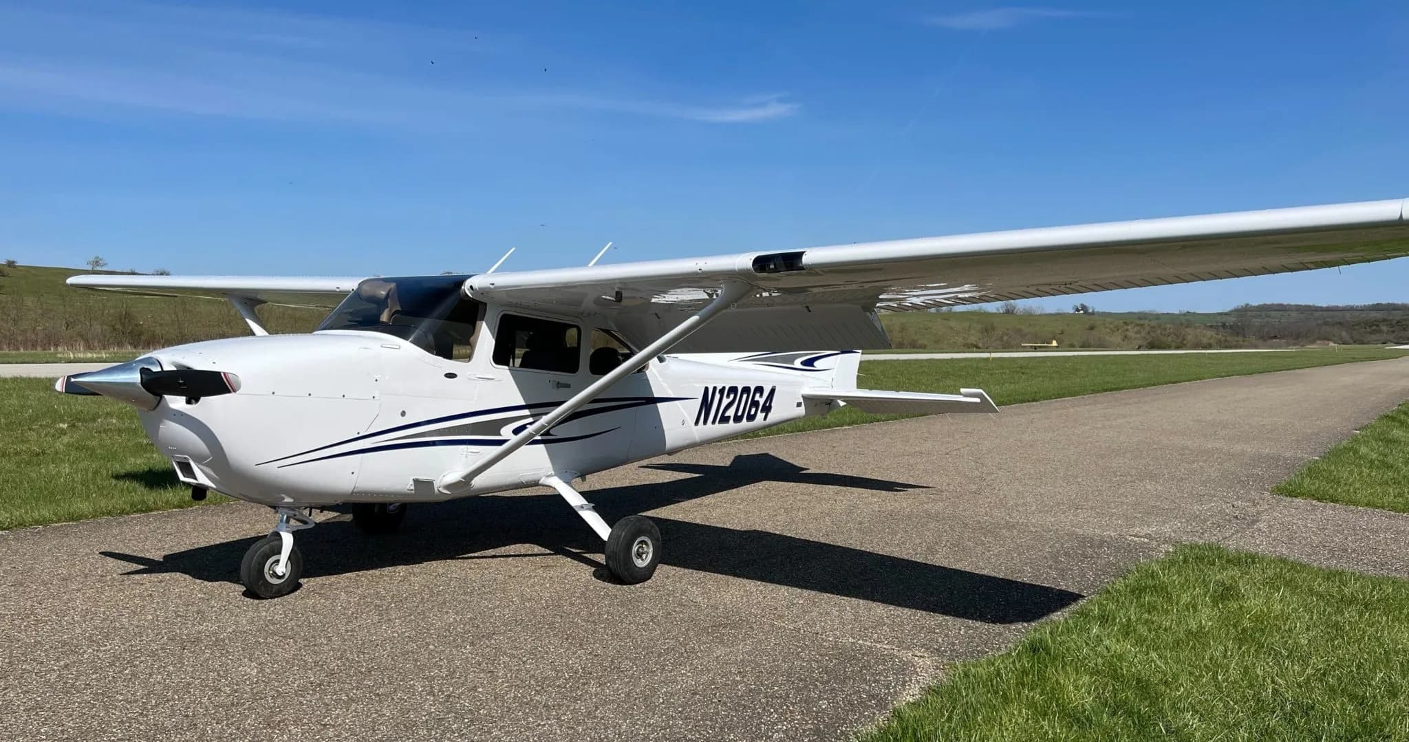 A white Cessna-style aircraft with blue and gray stripes is parked on an asphalt taxiway on a sunny day, with green hills in the background.