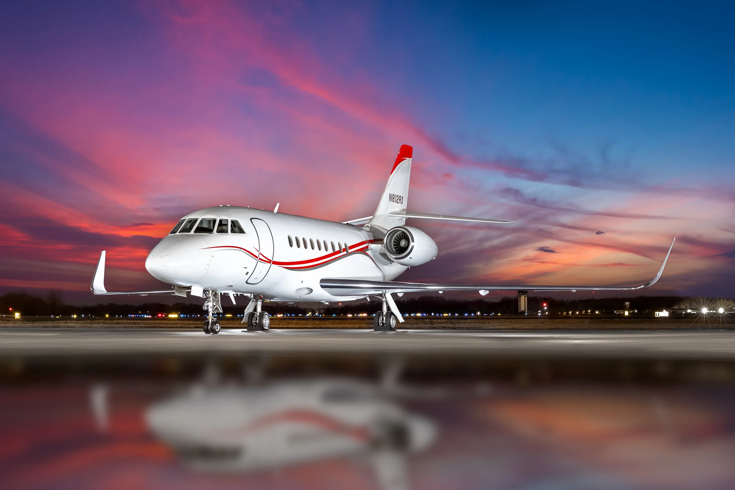 A white Dassault Falcon 2000LXS private jet with red and black stripes, registration N812RX, parked on a tarmac at sunset.