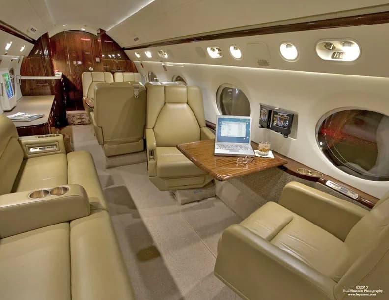 The aircraft's interior features tan leather seats, wood accents, and a laptop on a table.