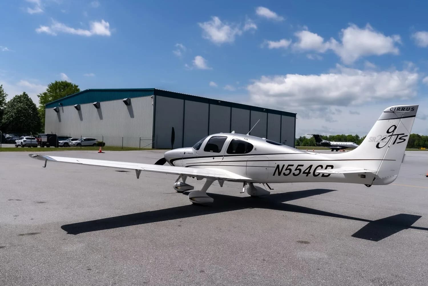 A rear three-quarter view of a silver and white Cirrus SR22T aircraft with registration N554CB parked on an airport tarmac next to a hangar.