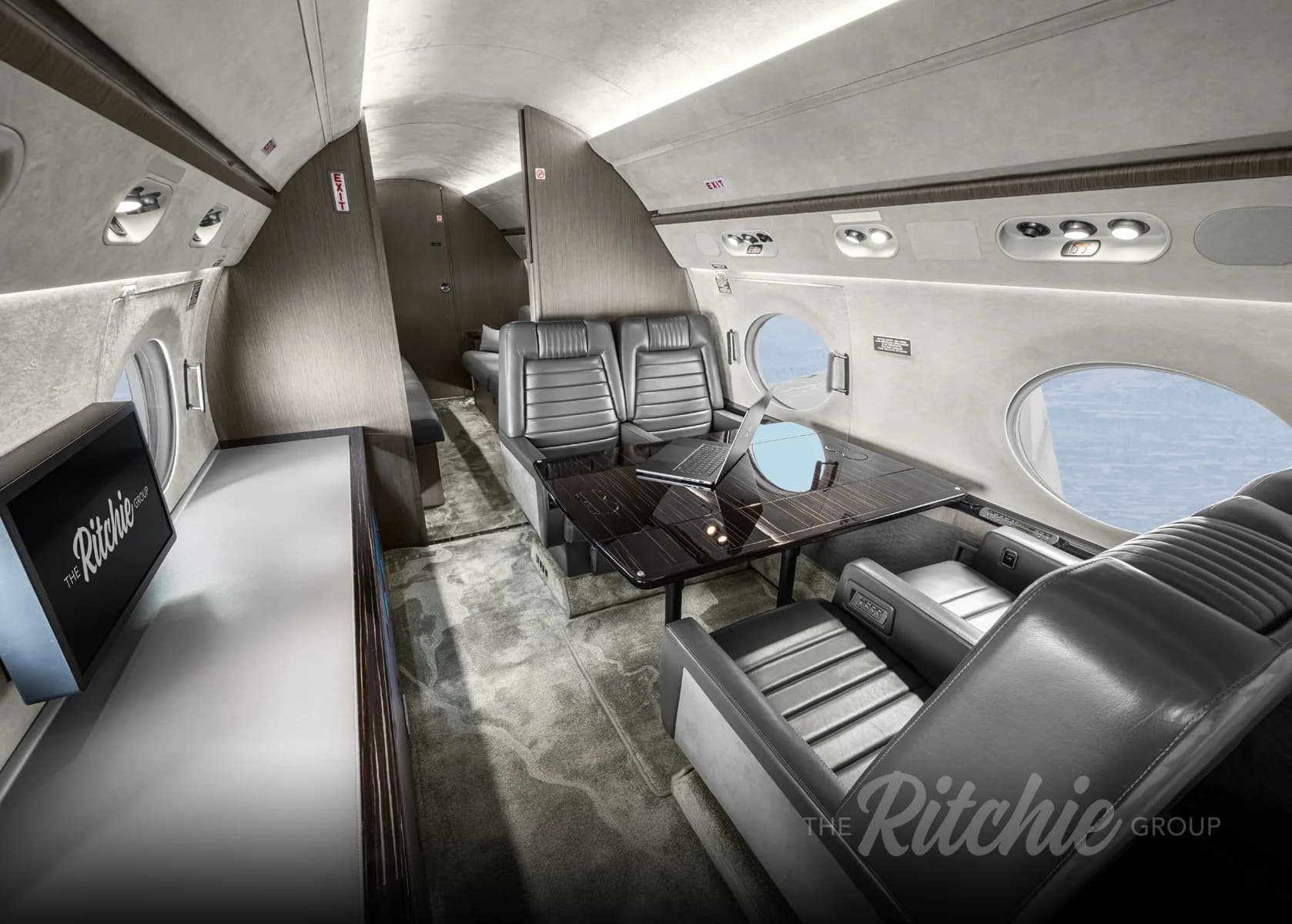 A view inside the Gulfstream G650 cabin, showing grey leather seating, a dark wood table with a laptop, and a monitor.