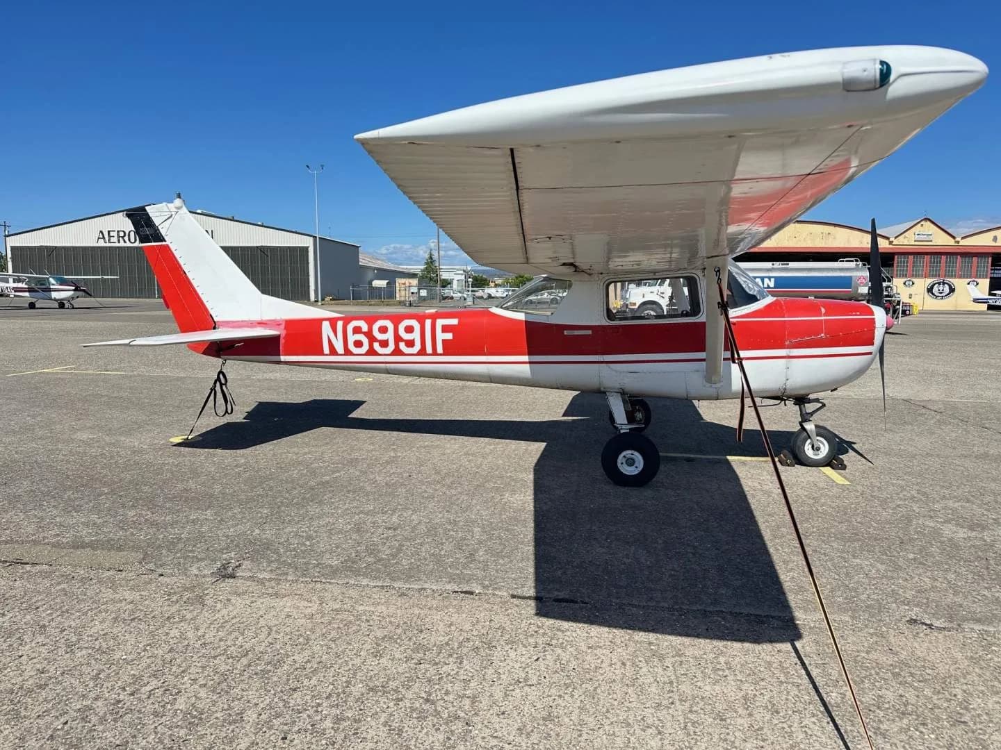 A red and white Cessna 150, tail number N6991F, parked on an asphalt apron with hangars in the background.