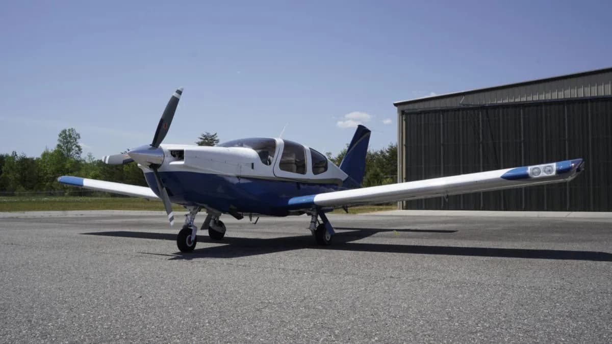 A front-left view of a white and blue single-engine aircraft with a propeller on an airfield.