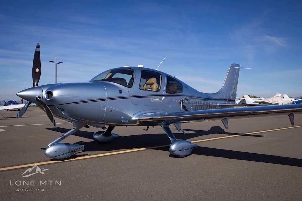 Front-left view of a silver Cirrus SR22 aircraft, N512HW, parked on an airfield under a blue sky.