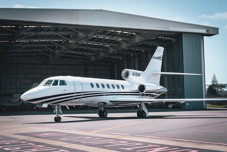 A white and black Falcon 7X aircraft is parked in front of an open hangar.