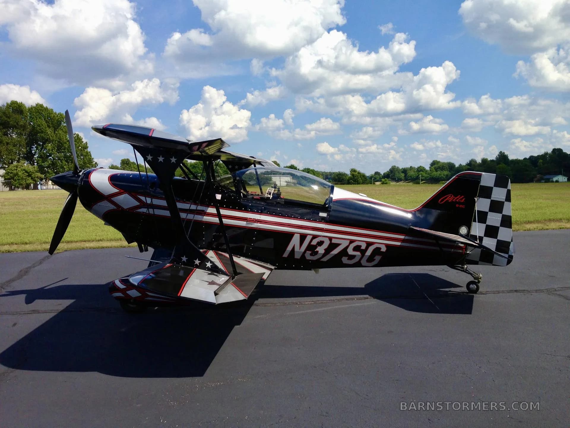 A black Pitts biplane with red and white stripes and stars, and a checkered tail, parked on an asphalt runway under a partly cloudy sky.