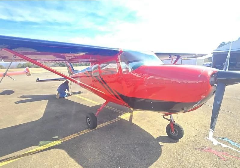 A red and black single-engine propeller aircraft is parked on an airfield with a person kneeling near the tail.