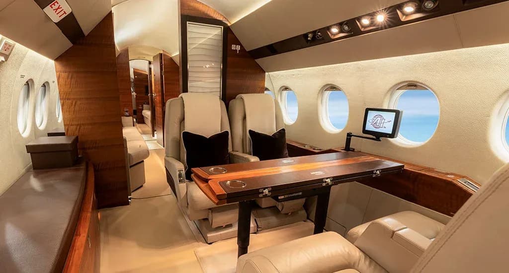 A section of a business jet's cabin with beige seats, a fold-out wooden table, and large windows.