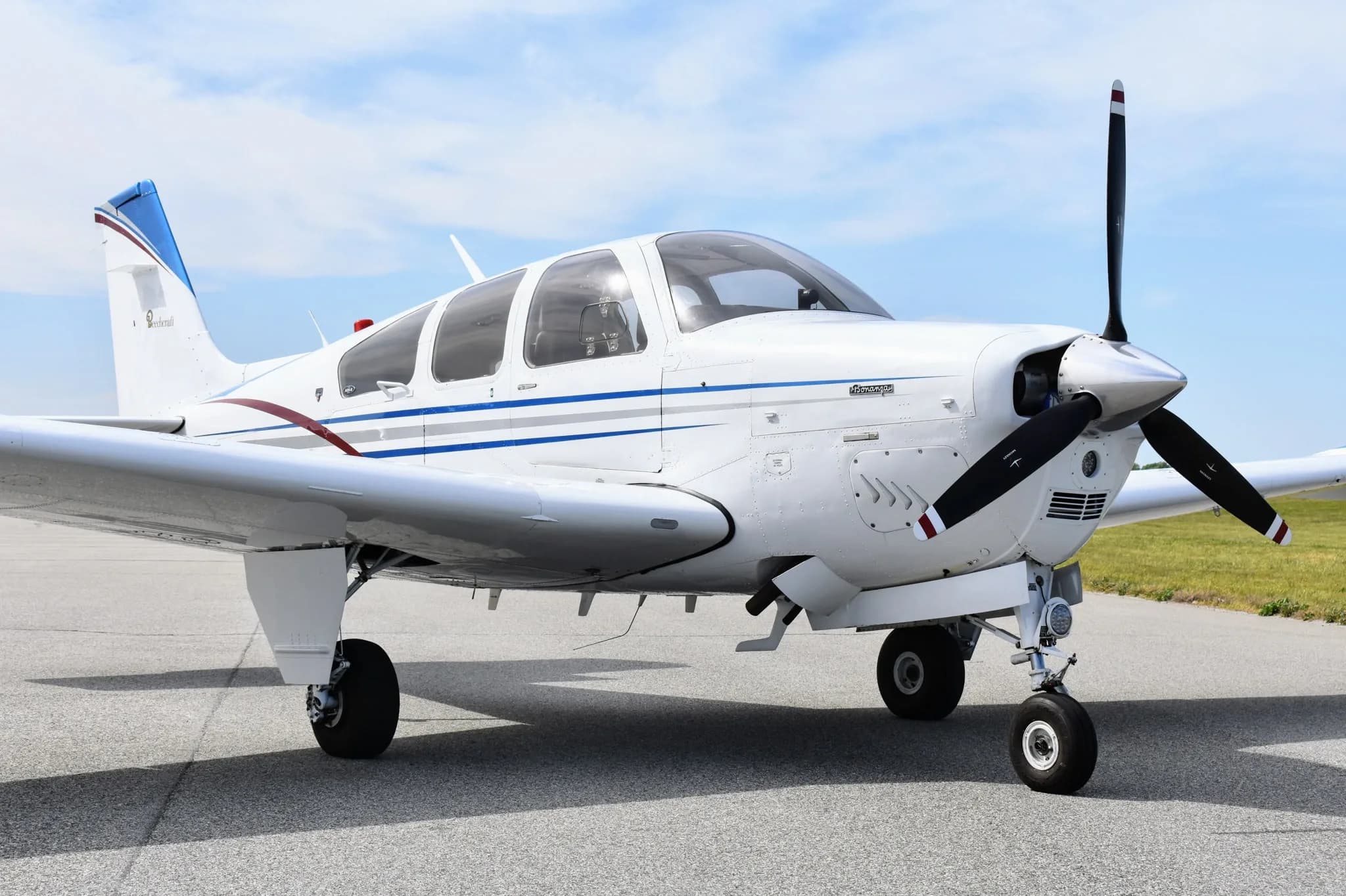 A close-up front view of a white Beechcraft Bonanza F33A with blue and maroon stripes, parked on an asphalt tarmac.
