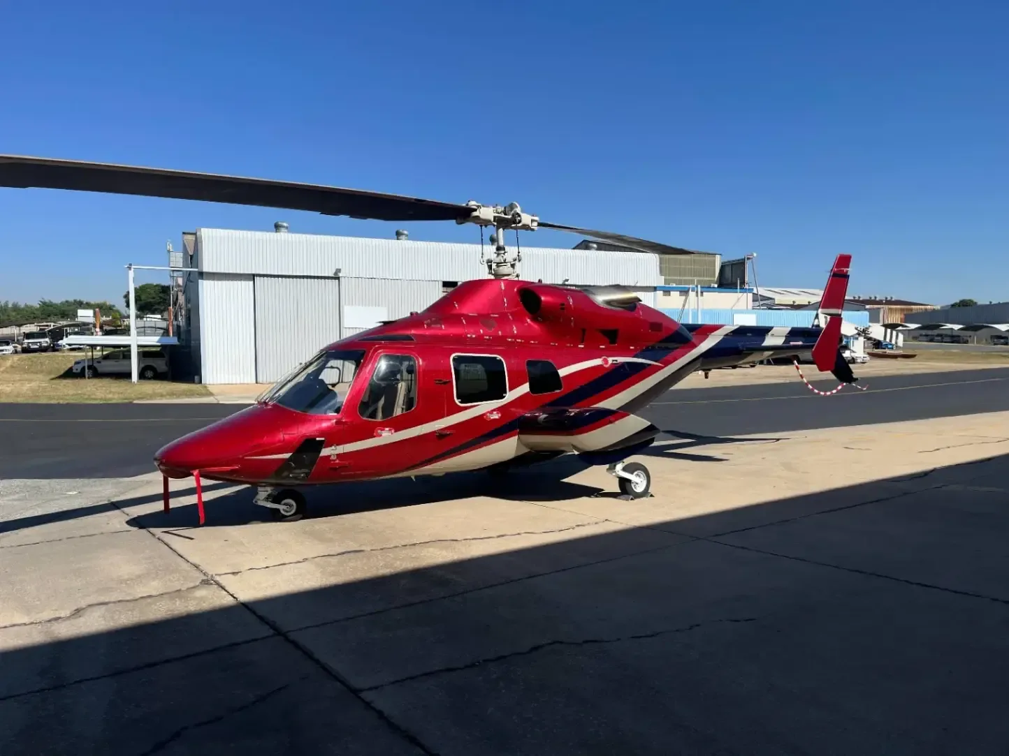 A red and white helicopter sits on the tarmac in front of a hangar.