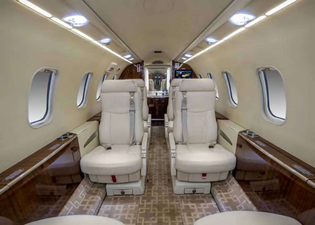 The luxurious interior cabin of a private jet, featuring light-colored leather seats, wood accents, and overhead lighting.