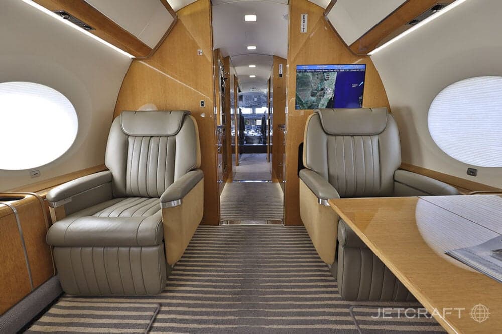 A view down the aisle of a business jet cabin with light brown leather seats, wood paneling, and a screen displaying a map.