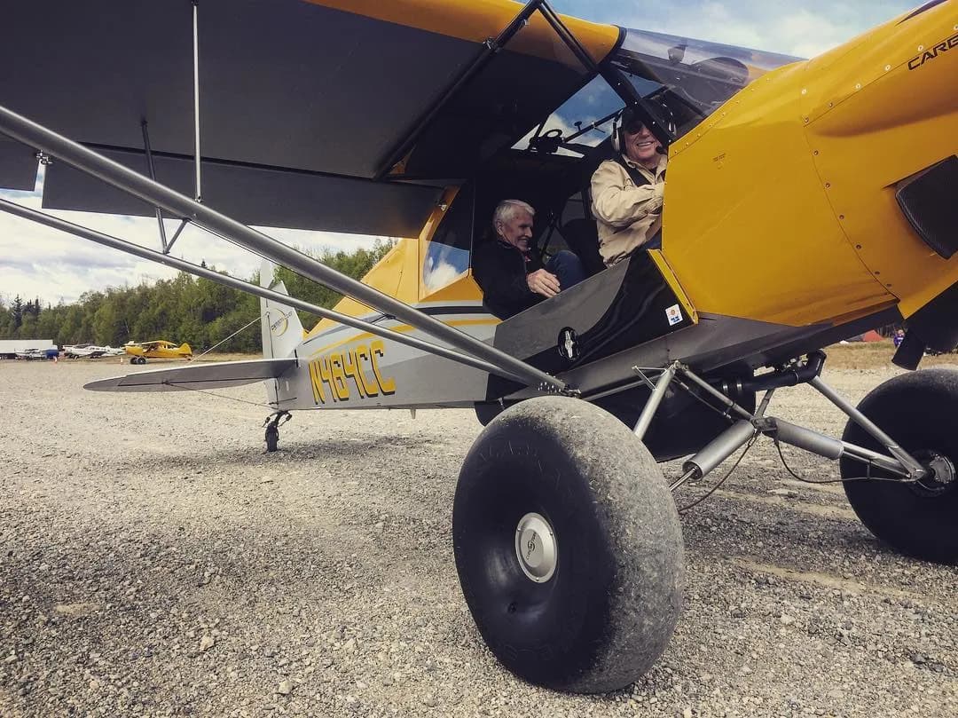 A yellow and grey Carbon Cub EX-2 (N464CC) with large tundra tires is on a gravel airstrip, with two men in the cockpit.