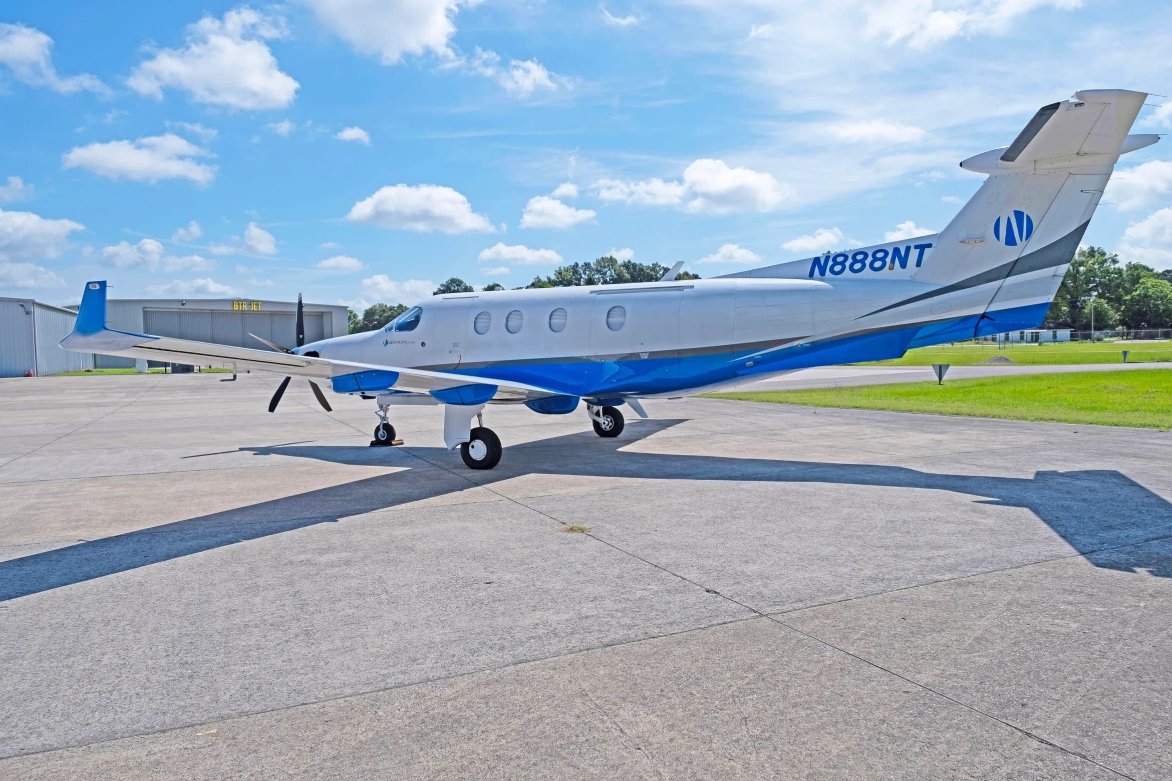 A white and blue Pilatus PC-12 aircraft, tail number N888NT, parked on a tarmac with hangars in the background.