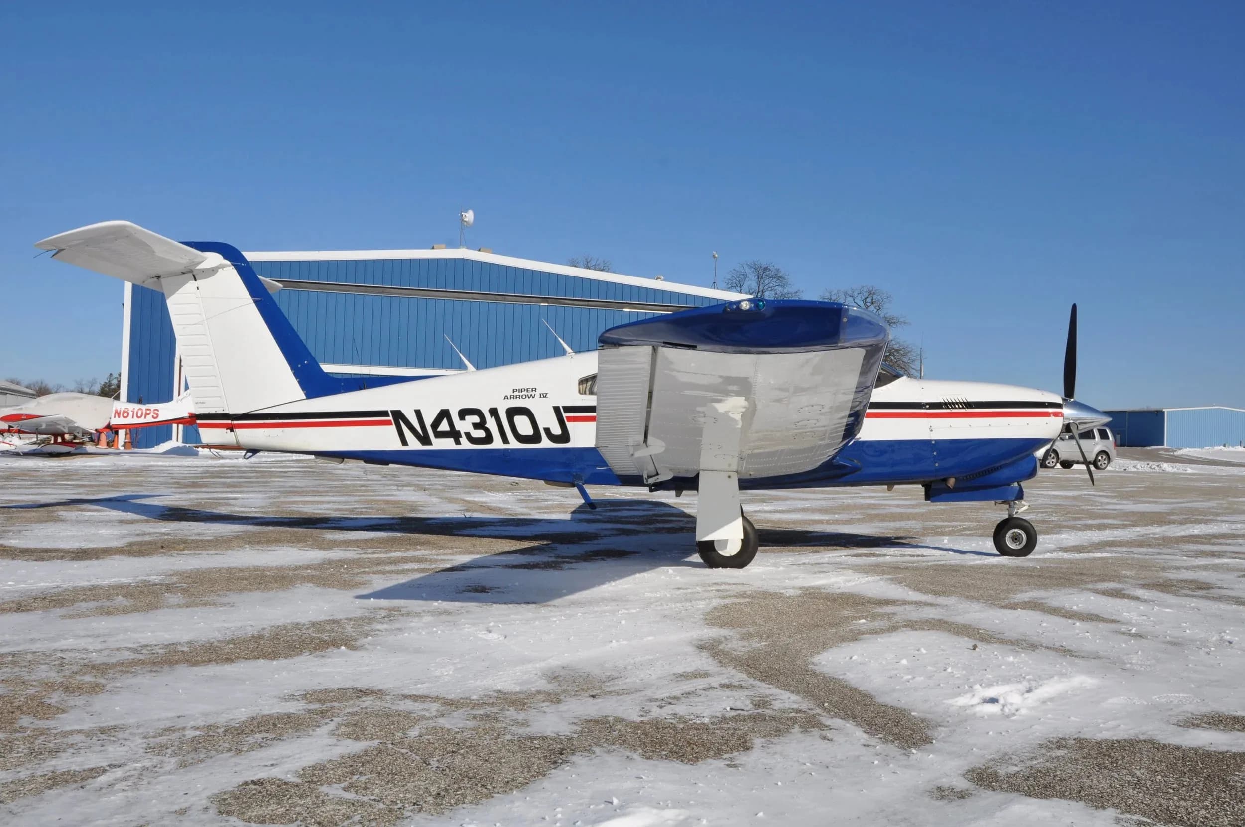 A white and blue Piper Arrow IV aircraft with registration N4310J parked on a snowy tarmac, viewed from the front-right side.