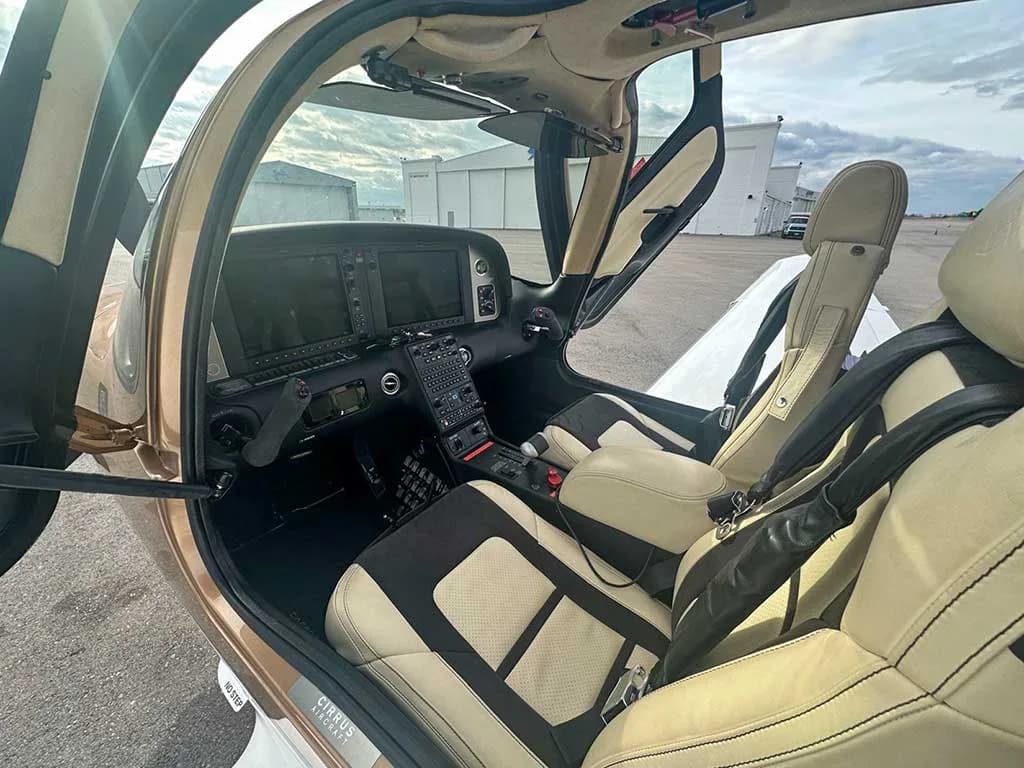 The cockpit of a Cirrus SR22 aircraft, showing the instrument panel and two-tone beige and black front seats with open doors.