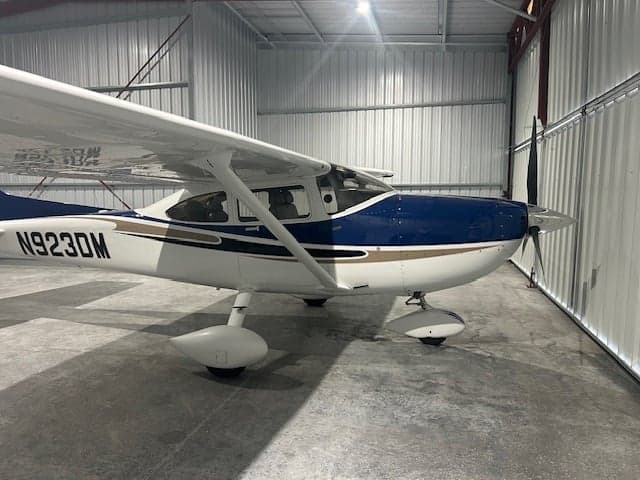 A white and blue single-engine propeller aircraft, N923DM, parked inside a metal hangar, viewed from the right side.