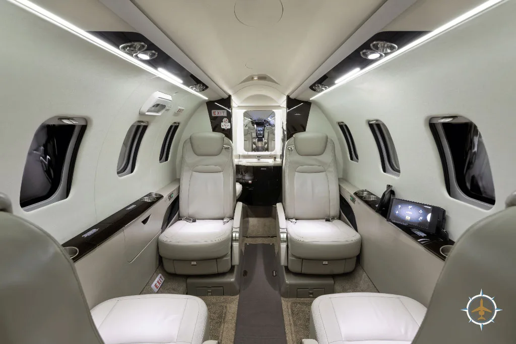 A view down the aisle of a business jet cabin, showing light-colored leather seats and a table with a magazine.