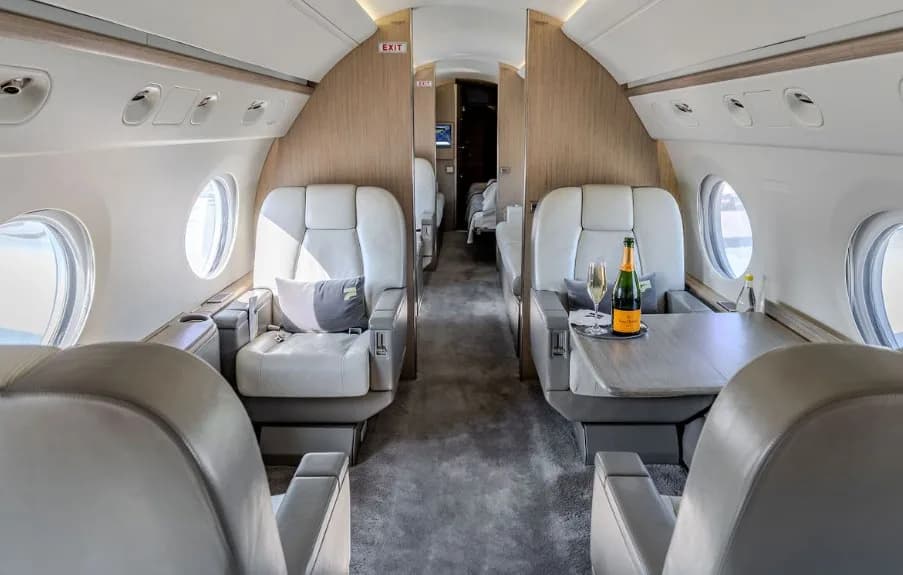 The luxurious interior cabin of a business jet, featuring light grey leather seats, a table with champagne, and wood paneling.