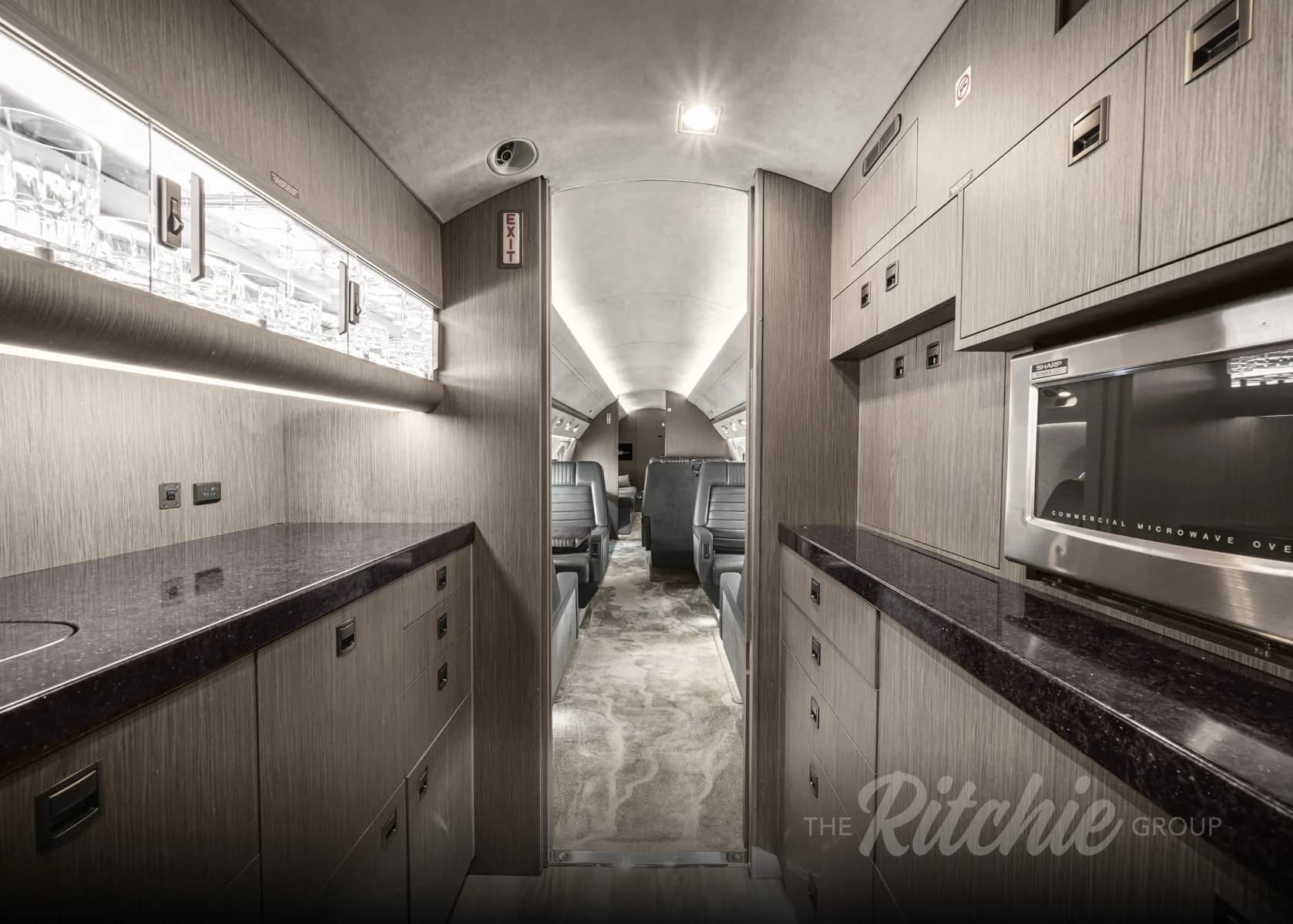 The galley area of the Gulfstream G650, featuring dark wood cabinetry, a microwave, and a view into the main cabin.