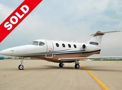 A white and brown business jet, registration N855RM, parked on an airfield under a cloudy sky.