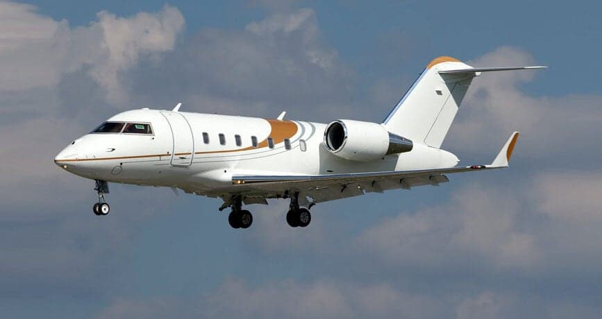 A white and gold Bombardier Challenger aircraft is shown in flight with landing gear down.