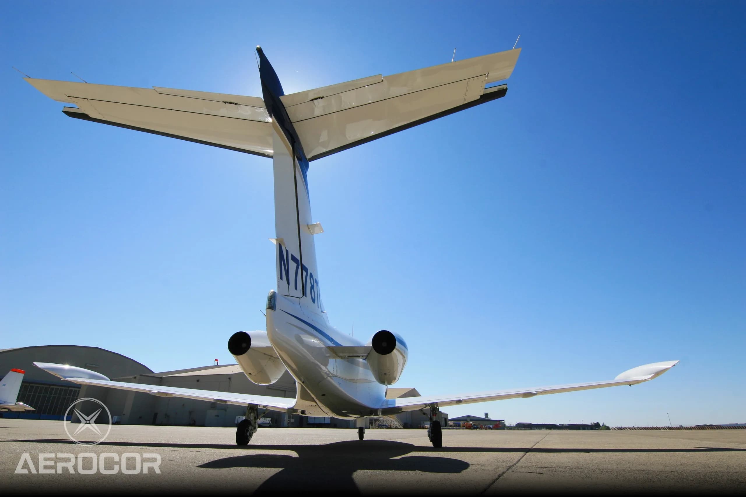 A rear view of a white Eclipse 500 jet with blue and gold stripes on the tarmac, silhouetted against a bright blue sky.