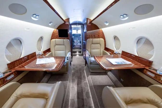 The interior cabin of a private jet, showing two sets of facing light-colored leather seats with polished wood tables between them.