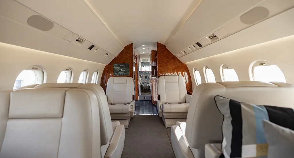 The spacious cabin interior of a business jet, showing rows of light-colored seats and a view towards the cockpit.