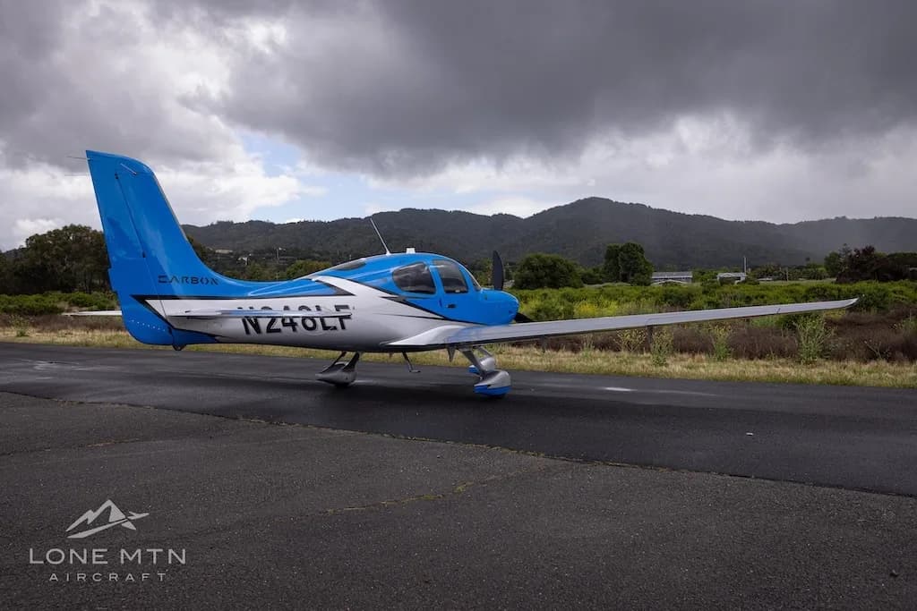A rear-side view of a blue and silver single-engine propeller aircraft (Cirrus SR22) with registration N248LF parked on a wet tarmac under a cloudy sky.