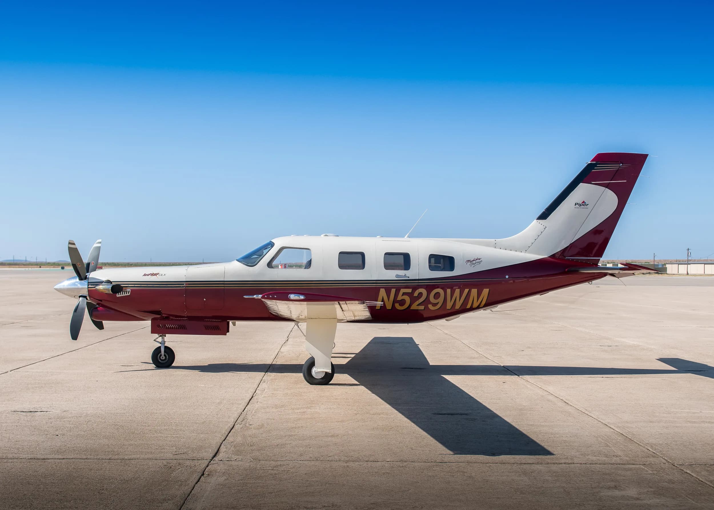 A side view of a maroon and white Piper M350 aircraft (N529WM) parked on an asphalt ramp under a clear blue sky.