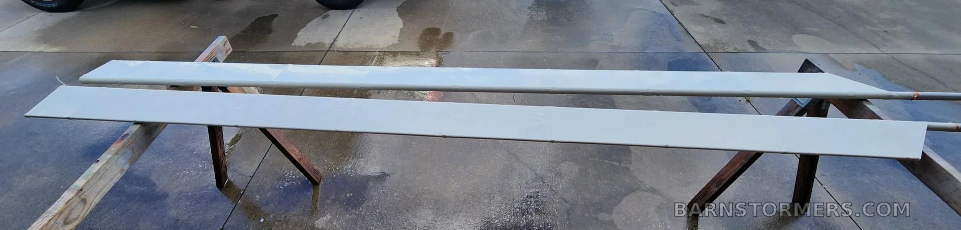 Two long, flat light grey aircraft components, possibly control surfaces, resting on sawhorses on a wet concrete pad.