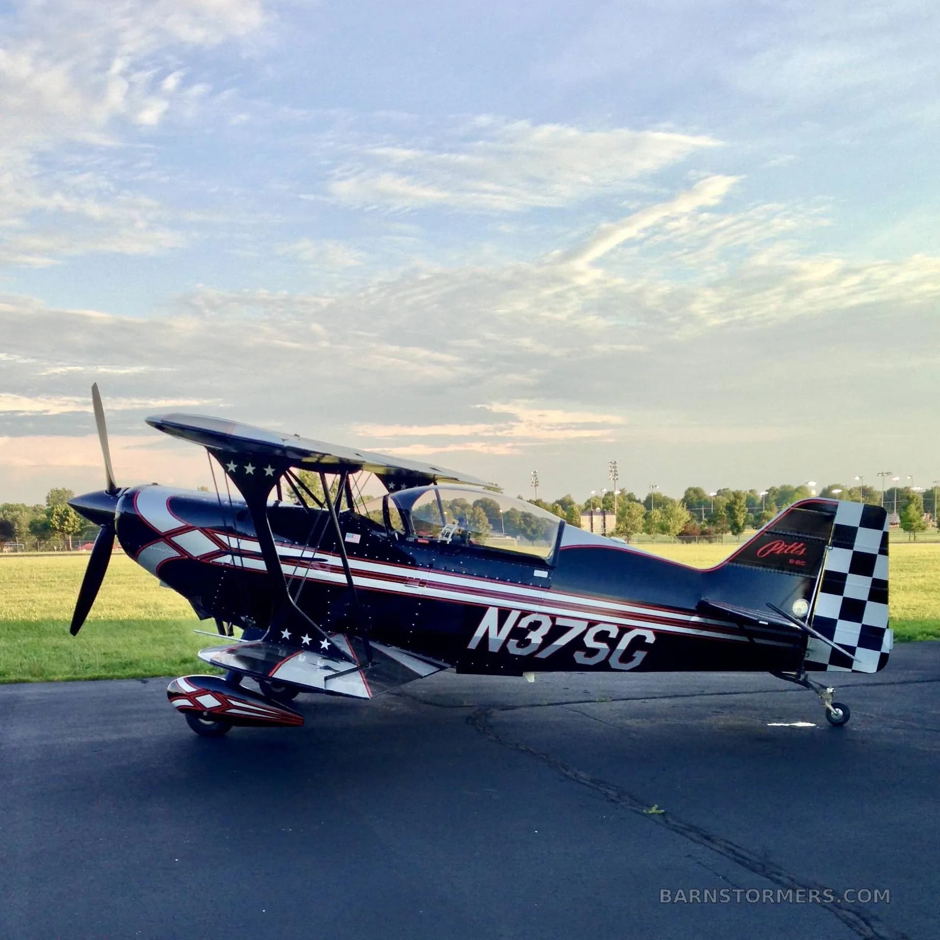 A black Pitts biplane with red and white stripes and stars, and a checkered tail, parked on an asphalt runway at dusk.