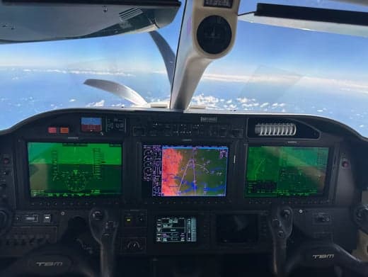 The cockpit of a TBM aircraft showing multiple glass displays and the view of clouds and ocean from the windshield.