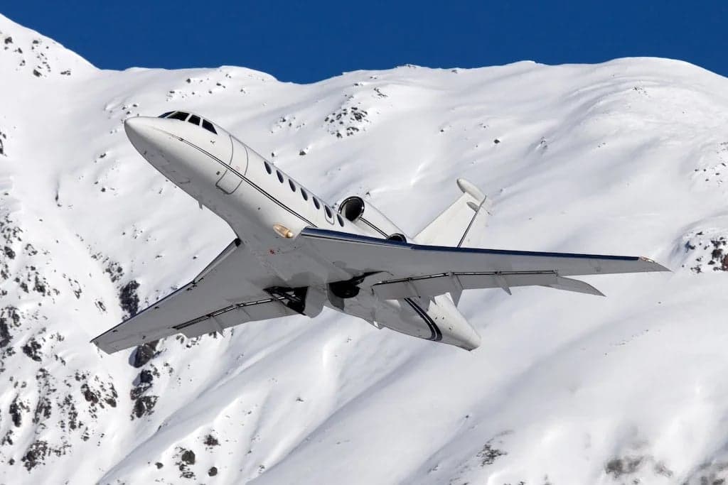 A white Falcon jet climbs over a snowy mountain range.