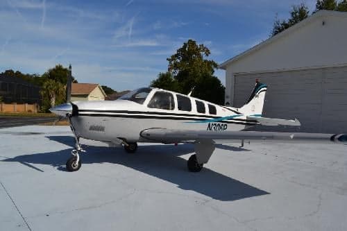 A Beechcraft Bonanza aircraft with a white and blue paint scheme parked outside a hangar.
