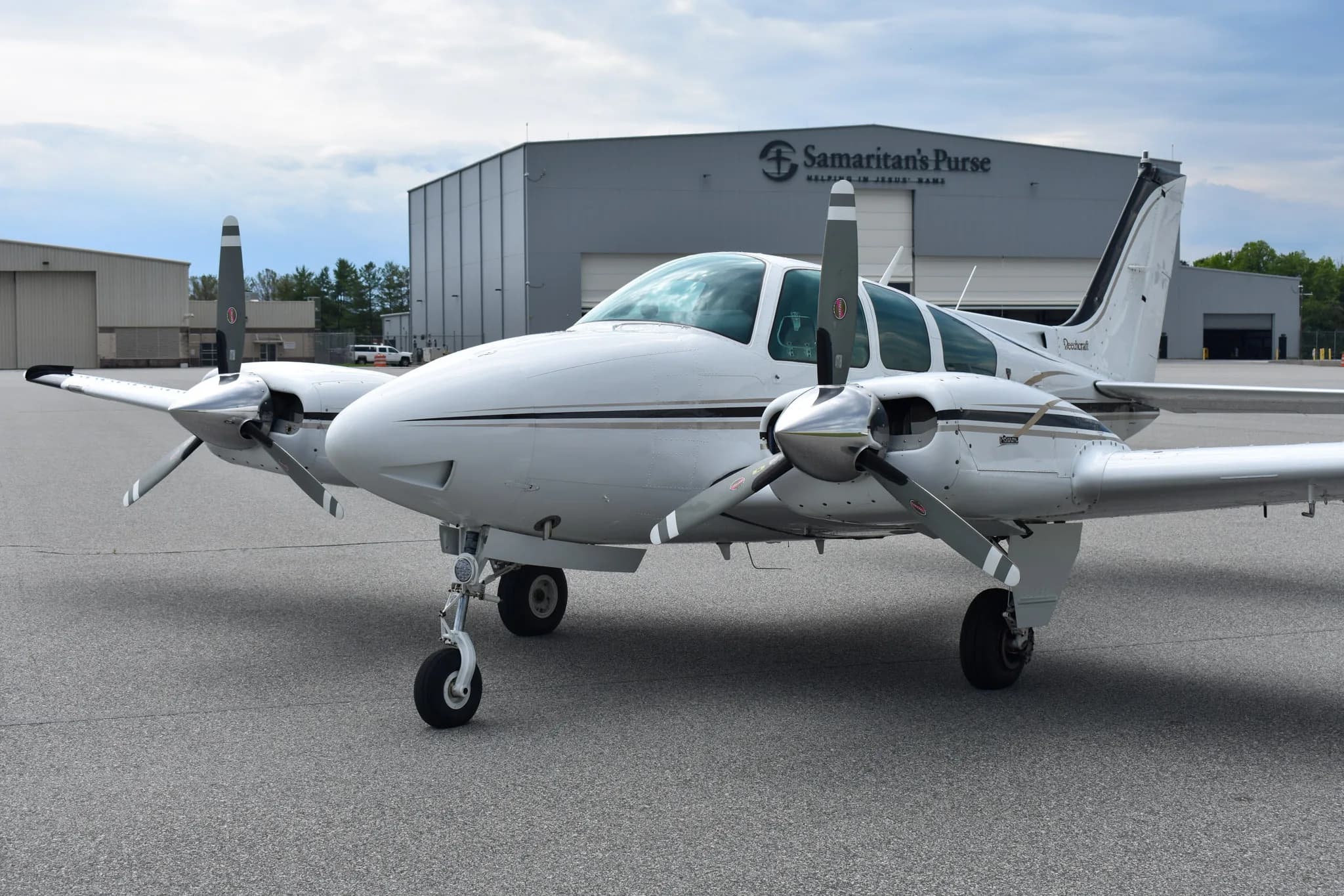 A front view of a white and black Beechcraft Baron aircraft, N6747V, parked on an asphalt runway in front of a 'Samaritan's Purse' hangar.