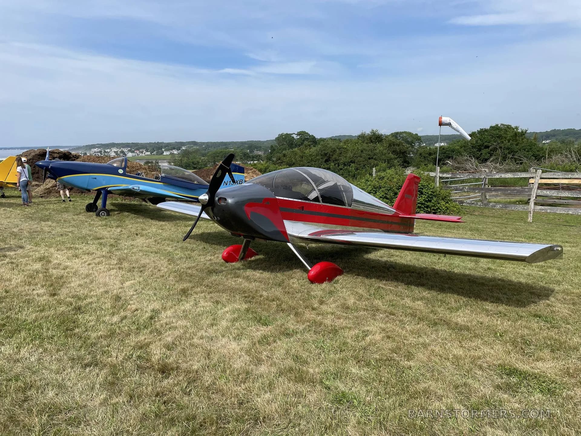 A red and silver experimental aircraft is parked on a grassy field next to a blue aircraft, with a windsock in the background.