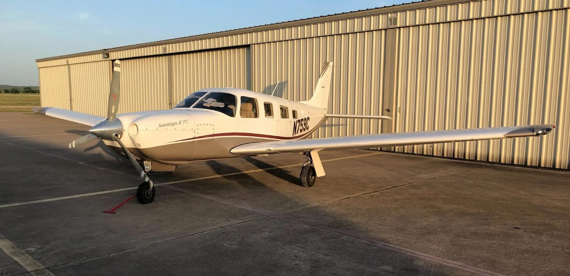 A white and tan Piper Saratoga II TC aircraft (N759C) parked on an asphalt ramp in front of a hangar at sunset.