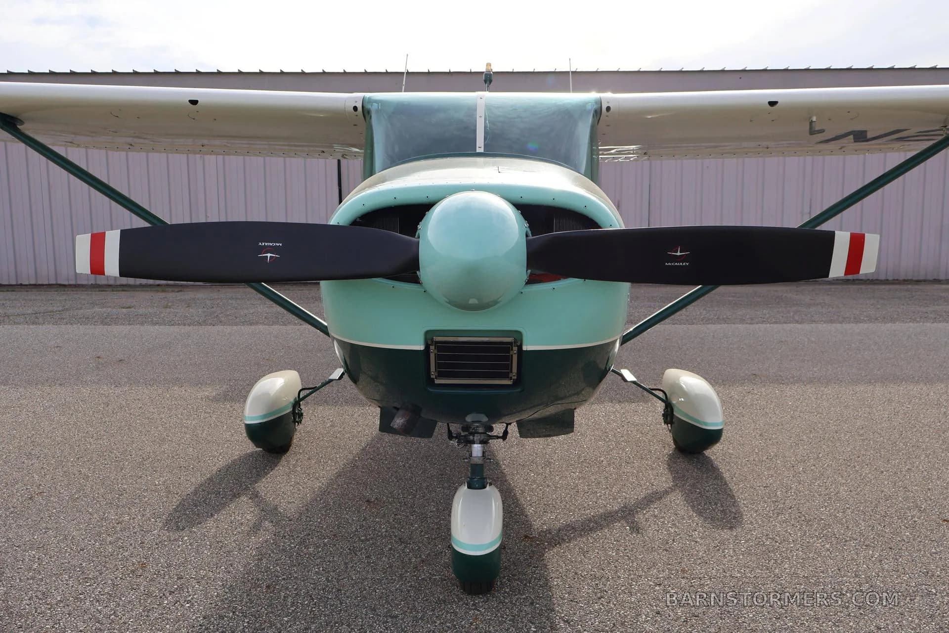 A green and white single-engine propeller aircraft (Cessna 172) is viewed head-on, showing its propeller and landing gear.