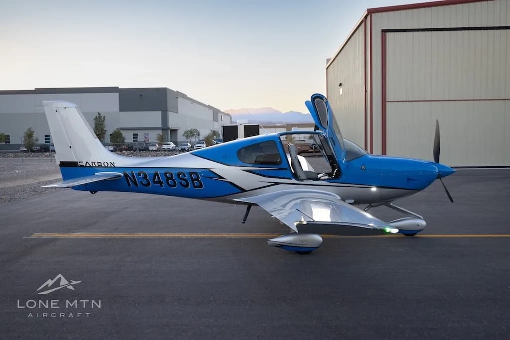 A side view of a blue and silver Cirrus SR22 aircraft with open doors parked on an airfield with buildings in the background.