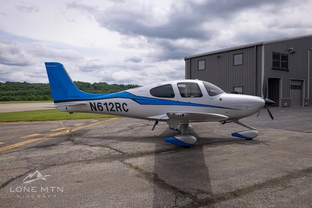 A silver and blue Cirrus SR22T Carbon aircraft, registration N612RC, parked on an airfield next to a hangar.
