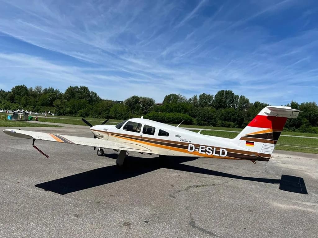A side view of a white and brown Piper Arrow III (D-ESLD) parked on an airfield under a blue sky.